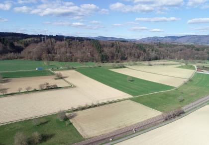 Hochrheintal westlich von Schwoerstadt – Blick zum Dinkelberg Aus großer Höhe blickt man über flache, rechteckig angelegte Felder und Wiesen zwischen einer Bahntrasse unten und einer Autostraße in der Bildmitte. Zum Hintergrund hin erheben sich ausgedehnte Waldhänge.