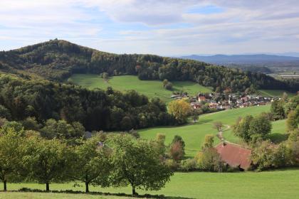 Blick aus südwestlicher Richtung über Waldkirch-Suggental Weiter Blick von erhöhtem Standort über eine hügelige grüne Landschaft mit Wiesen und Bäumen. Im Hintergrund, nach links hin ansteigend, erstreckt sich ein bewaldeter Bergrücken. Rechts liegt in einer Talmulde eine Siedlung.