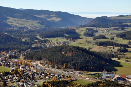 Zwischen Titisee und Höllental Von weit oben blickt man auf eine größere Ortschaft links unten sowie eine weite, vorwiegend grüne und hügelige Landschaft, die bis zum Hintergrund von Waldrücken durchsetzt ist. Hinten rechts folgen dann noch bewaldete Berge.