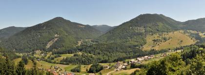 Der Präger Gletscherkessel Panoramabild zweier hoher, nebeneinander liegender bewaldeter Berge. Rechts unten sowie links vorn verteilen sich Häuser mit Walmdächern.