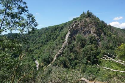 Fels im St. Blasien-Granit mit vorspringendem Gang aus Granitporphyr auf der Westseite des Schlüchttals unterhalb der Burgruine Schnörringen Blick aus großer Höhe auf einen zum Hintergrund hin aufsteigenden, bewaldeten Bergkegel. Der obere Teil des Kegels zeigt Felsformationen, zudem verläuft auf der linken Seite, in der Bildmitte, ein vorspringender, mauerartiger Felsengang.