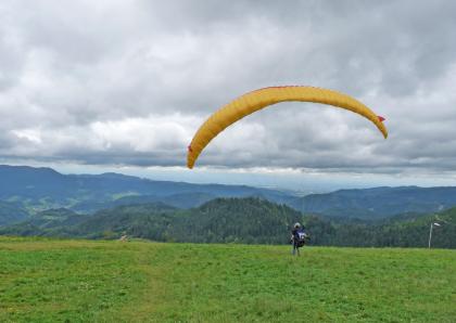 Blick vom Roßbühl nach Westen über das Renchtal zur Oberrheinebene bei Oberkirch Das Bild zeigt eine erhöht liegende Wiese mit Blick auf tiefer gelegene Nadelwälder, bewaldete Bergrücken sowie eine ferne Ebene. Rechts wartet ein Gleitschirmflieger auf seinen Absprung.