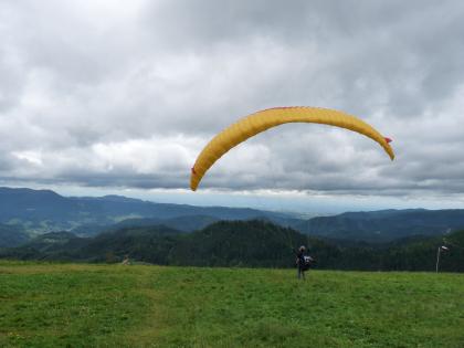Blick vom Roßbühl nach Westen über das Renchtal zur Oberrheinebene bei Oberkirch Das Bild zeigt eine erhöht liegende Wiese mit Blick auf tiefer gelegene Nadelwälder, bewaldete Bergrücken sowie eine ferne Ebene. Rechts wartet ein Gleitschirmflieger auf seinen Absprung.