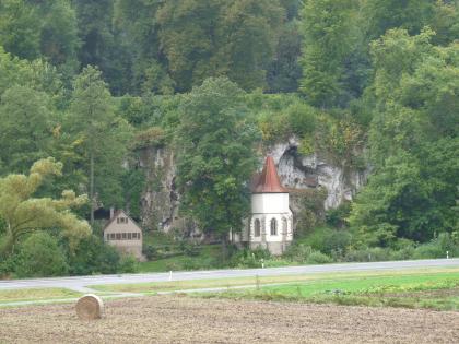 Kalktufffelsen im Jagsttal bei St. Wendel zum Stein Hinter der Kapelle St. Wendel ragt eine grauweiße, zerfurchte Felswand empor. Teilweise ist das Gestein von dichtem Buschwerk überwuchert; rechts und links umrahmen es hohe Bäume. Rechts ist außerdem eine Höhle zu erkennen.