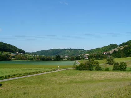 Blick das Kochertal entlang nach Westen in Richtung Ernsbach Das Bild zeigt eine weite, grüne Landschaft, die von schmalen Straßen und Bäumen durchzogen ist. Im Hintergrund erheben sich von links über die Mitte bis zum rechten Rand bewaldete Anhöhen.