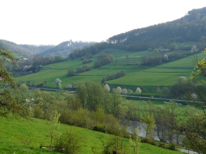 Kochertal bei Braunsbach-Steinkirchen, im Hintergrund Schloss Tierberg Über einem nur halb sichtbaren, von Bäumen gesäumten Fluss zieht sich rechts ein grüner, oben bewaldeter Hang hinauf. Straßen und Wege durchziehen ihn. Im Hintergrund zeigen sich weitere bewaldete Erhebungen. Auf einer davon thront ein altes Schloss.
