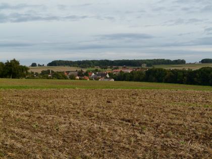 Raboldshausen liegt in einem Muldental bereits im Muschelkalk am Übergang zum Lettenkeuper Eine braune, mit Stoppeln bedeckte Ackerfläche erstreckt sich bis zur Bildmitte, gefolgt von einem weiteren, grünen Acker. Dahinter verschwinden Bäume und eine kleine Ortschaft in einer tiefen Mulde. Zum Horizont hin steigen wieder Äcker und Wald empor.
