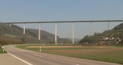 Das Kochertal bei Geislingen am Kocher Eine hohe, von schmalen Pfeilern getragene Autobahnbrücke überwindet eine Talaue zwischen zwei bewaldeten Berghängen. Die flache Aue wird von einer kurvigen Straße eingefasst. Am rechten Berghang verteilen sich Häuser.