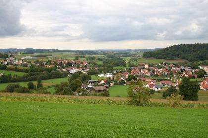 Blick über Bühlerzell nach Norden Hinter bepflanzten Ackerflächen im Vordergrund breitet sich eine weite, ebene Landschaft mit Siedlungen und Waldinseln aus. Rechts schiebt sich eine größere Waldfläche ins Bild; links zeigen sich grüne Hügel. Im Hintergrund sind flache Höhen erkennbar.