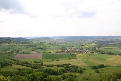 Der südliche Teil der Haller Stufenrandbucht Blick aus großer Höhe über eine weite, hügelige Landschaft mit Feldern, Wiesen, Bäumen, Waldstreifen und kleineren Siedlungen. Im Hintergrund erheben sich flache bis abgerundete, bewaldete Berg- und Hügelkämme.