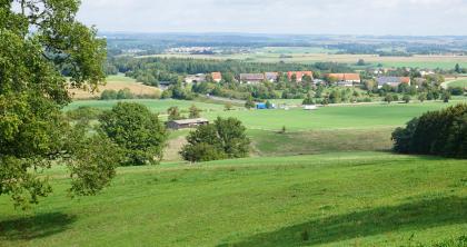 Blick vom Albanstieg bei Oberalfingen nach Nordosten über den Hangfuß im Opalinuston mit dem Ort Immenhofen auf die weiter hinten gelegenen, von Goldshöfe-Sanden bedeckten Unterjura-Flächen. Blick von einem erhöht liegenden, zur Bildmitte hin abfallenden Wiesenhang auf eine flachwellige Landschaft mit Wiesen, Äckern, Waldstreifen sowie einer kleinen Ortschaft. Im Hintergrund sind langgestreckte waldige Höhen erkennbar.