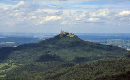 Blick vom Trauf der Mittleren Alb nach Norden über die auf dem Ausliegerberg Zoller gelegene Burg Hohenzollern Ein schönes Bild, das einen bewaldeten, zur Mitte hin ansteigenden Berg zeigt, bekrönt von einer stattlichen Burg. Im Vordergrund dehnen sich weitere Hänge aus, während der Hintergrund bis auf ferne Höhenzüge flach erscheint.