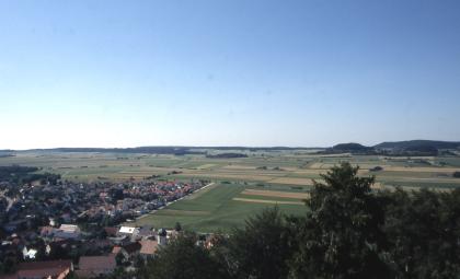 Blick von der Burgruine Hohenringingen auf das „Heufeld“ nördlich von Burladingen-Ringingen Über Baumwipfel rechts blickt man über eine weite, landwirtschaftlich genutzte Ebene sowie eine Ortschaft links. Im Hintergrund rechts sind unter wolkenlosem Himmel bewaldete Hügel erkennbar.