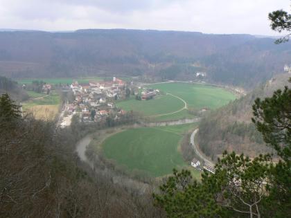 Blick auf Beuron von Südosten Von erhöhtem Standpunkt aus blickt man auf eine flache, teils besiedelte und teils grüne Talaue, die ein Fluss C-förmig durchquert. Steile, bewaldete Höhenzüge umrahmen die Aue. Im Zentrum der Siedlung liegt ein Kloster.
