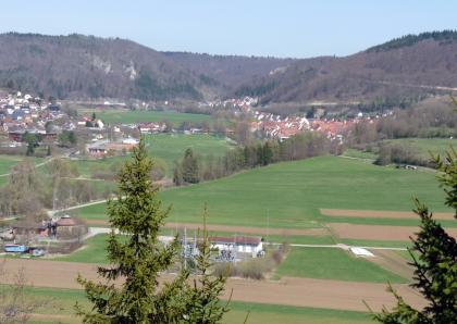 Talwindungen der Donau bei Fridingen – Blick vom Bergsteig nach Norden Von erhöhtem Standpunkt aus blickt man eine ebene Acker- und Grünlandschaft, die links und im Mittelgrund besiedelt ist. Straßen, Wege und Baumstreifen durchqueren die Ebene, mehrere bewaldete Höhenzüge begrenzen sie im Hintergrund.