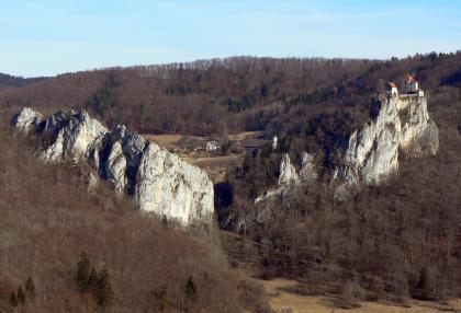 Massenkalkfelsen mit dahinter liegender Zementmergelschüssel bei Schloss Bronnen östlich von Fridingen an der Donau Das Bild zeigt zwei sich gegenüber liegende, aus bewaldeten Hängen aufsteigende, weiß und grau gefärbte Felstürme. Auf dem Felsen rechts thront eine noch erhaltene Burg. Im Hintergrund zeigt sich eine Ebene sowie ein weiterer bewaldeter Höhenzug.
