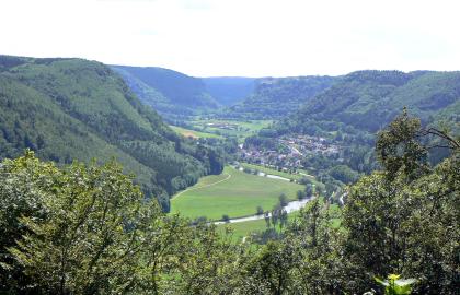 Das obere Donautal bei Beuron-Hausen im Tal Von hohen bewaldeten Steilhängen umschlossen, liegt eine breite grüne Ebene in der Bildmitte. Ein im Bogen verlaufender Fluss durchquert die Ebene, die sich zum Hintergrund hin fortsetzt. Eine Siedlung liegt am Fuß der Anhöhe rechts.