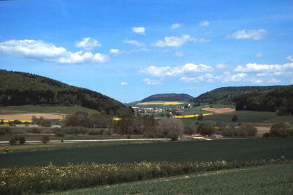Blick über das Aitrachtal nach Norden zum Fürstenberg Das Bild zeigt eine grüne Mischlandschaft mit Baumgruppen im Mittelgrund und bewaldeten Hängen links und rechts. In der Mitte liegt weiter entfernt eine dritte Anhöhe, die am Fuß besiedelt ist.