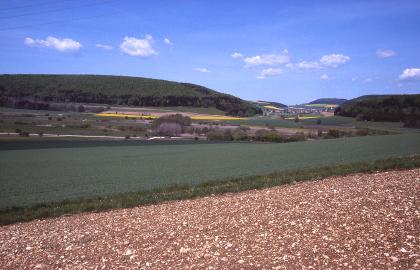 Blick über das Aitrachtal nach Nordnordwesten An einen sehr steinigen Ackerboden im Vordergrund schließen sich dunkelgrüne Nutzflächen an. Im Hintergrund sind weitere Ackerflächen sowie links und rechts der Mitte ansteigende Höhenzüge erkennbar.