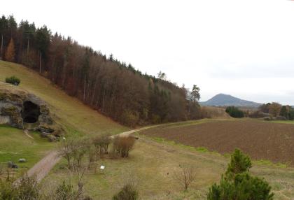 Brudertal östlich von Engen Links eines Weges steigt ein grüner, teilweise bewaldeter Hang auf. Auch der Eingang einer Felsenhöhle ist erkennbar. Rechts teilt sich das Gelände in Acker und trockene Wiese auf und fällt nach vorne ab. Im Hintergrund ist eine steile Erhebung zu sehen.