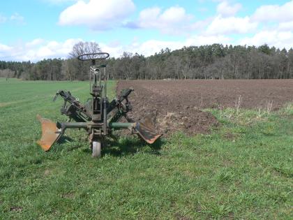 Rotbraune Ackerböden auf den Buntsandstein-Randplatten bei Bad Teinach-Zavelstein Das Bild zeigt ebene Grünlandflächen, an die sich rechts eine Ackerfläche mit rotbraunem Boden anschließt. Im Vordergrund ist ein ein alter Pflug zu sehen. Im Hintergrund steht Wald.