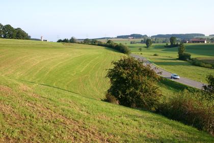 Das Mühlbachtal bei Bad-Wurzach-Eggmannsried (Lkr. Ravensburg) Von einer kurvigen Straße durchschnitten, steigen links und rechts flache grüne Hügel auf, im Vordergrund links mit Dürrespuren. Rechts sowie im Hintergrund werden die Hänge von Gebüsch und Bäumen begrenzt.