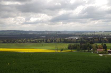 Blick von der Grabener Höhe in südöstliche Richtung in das Wurzacher Ried Unter einem von grauen Wolken durchzogenen Himmel breitet sich eine weite Landschaft mit Wiesen, Feldern und ausgedehnten Waldflächen aus. Im Hintergrund sind mehrere Felder zu sehen, auf denen Wasser steht.