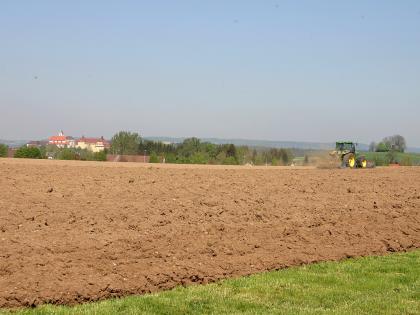 Frühjahrsbestellung im Jungmoränen-Hügelland in der Umgebung von Bad Waldsee Blick auf einen rötlich braunen, mit Steinen durchsetzten Acker, der von einem Traktor befahren wird. Im Hintergrund Bäume und Gebäude.