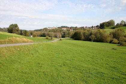 Aue der Wolfegger Ach bei Wolfegg Blick über eine mit Bäumen begrenzte Wiesenlandschaft, die nach links vorne abfällt, nach rechts und links oben jedoch ansteigt. Im Hintergrund rechts ist auf einem bewaldeten Rücken eine Kirche erkennbar.