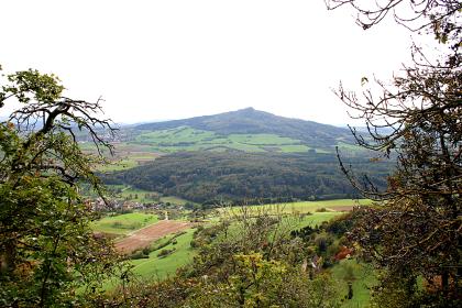 Blick auf den Hohenstoffeln Blick durch Bäume hindurch über eine Landschaft mit Feldern, Wiesen, Siedlungen und Waldstücken auf einen bewaldeten Vulkankegel.