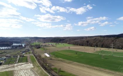 Hochrheintal beim Kraftwerk Ryburg-Schwörstadt – Blick rheinabwärts Aus großer Höhe blickt man über flache Wiesen und Felder, durchzogen von Bahnschienen und Autostraßen. Links im Bild ist ein breiter Fluß erkennbar; davor befindet sich ein Kraftwerk. Rechts breiten sich Wälder aus.