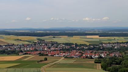 Blick über den Kraichgau nach Norden zum Katzenbuckel im Odenwald Weiter Blick aus großer Höhe über eine wellige Landschaft. Zwischen zahlreichen Äckern liegt eine Siedlung. Im Hintergrund sind Waldstreifen und bewaldete, langgezogene Berge zu erkennen.