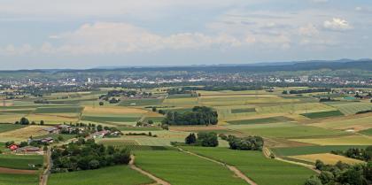 Aussicht vom Turm auf der Heuchelberger Warte nach Osten auf das Stadtzentrum von Heilbronn und die Keuperberge Weiter Blick aus großer Höhe auf eine flache bis wellige Landschaft mit zahlreichen Ackerflächen. Im Hintergrund, vor niedrigen Bergen mit bewaldeten Kuppen, liegt eine Stadt.