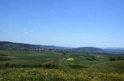 Am Fuß der Keuper-Schichtstufe bei Rottenburg am Neckar verzahnt sich die Gäulandschaft mit dem angrenzenden Keuperbergland Von einer blühenden Wiese geht der Blick talwärts zu einer bewirtschafteten, welligen Ebene mit grünen, braunen und gelben Ackerflächen. Im Hintergrund verteilen sich mehrere Höhenzüge unter einem blauen Himmel.