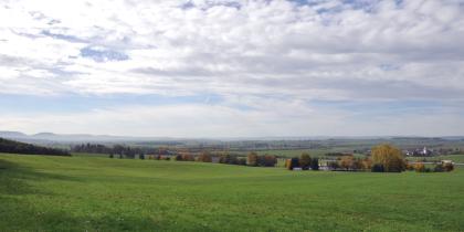 Blick vom Unterhang der Mitteljura-Schichtstufe bei Bad-Dürrheim-Sunthausen über die Baar-Hochmulde nach SSW Der Blick geht über weite, im Mittelgrund eingetiefte Grünflächen bis zu einer Autobahn. Dahinter ist die Landschaft flach und grün, mit Siedlungen und Wald. Ganz im Hintergrund sind verschiedene Hochflächen erkennbar.