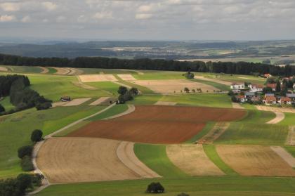 Mitteljura-Plateau bei Hausen ob Verena Von oben blickt man auf ein geschwungenes Bodenmosaik aus Grünland und Äckern. Zwei rötlich braune Äcker im Mittelgrund ragen dabei heraus. Im Hintergrund, nach einem Waldstreifen, sind links abgestufte Höhenzüge erkennbar.