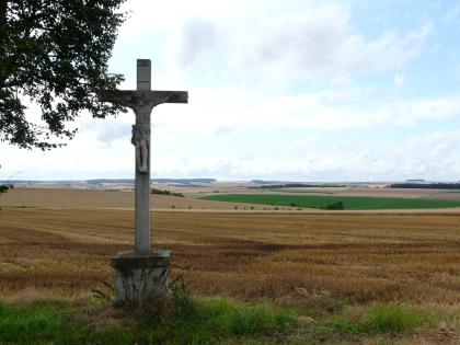 Flachwellige Gäulandschaft östlich von Tauberbischofsheim Von einem Wegkreuz links blickt man auf einen braunen, abgeernteten flachen Acker, der bis zum Mittelgrund reicht. Dahinter schließen sich weitere, nun leicht wellige Ackerflächen an.