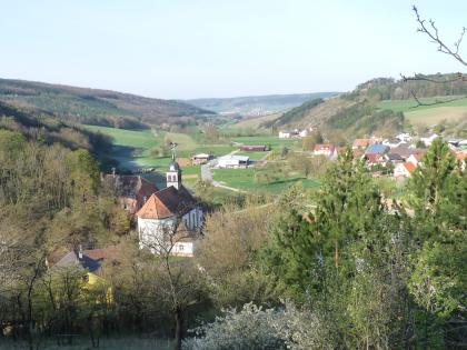 Welzbachtal bei Werbach-Werbachhausen Von erhöhtem Standort aus blickt man auf ein schmales, an den seitlichen Rändern hügeliges und bewaldetes Tal. Eine kleine Ortschaft mit Kirche verteilt sich links und rechts darin.
