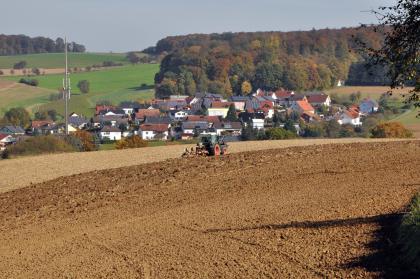 Blick nach Nordosten über das Schefflenztal bei Billigheim-Allfeld am Südwestrand des Baulands Das Bild zeigt im Vordergrund von einem Traktor gepflügte, rötlich braune Ackerflächen. Hinter einer angrenzenden Siedlung erhebt sich rechts ein bewaldeter, links von weiteren Äckern gebildeter Hang.