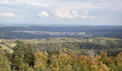 Am Nordrand des Buntsandstein-Schwarzwalds geht der Blick über die auf der Hochfläche zwischen Nagold- und Würmtal gelegene Ortschaft Huchenfeld nach Osten Blick aus großer Höhe über Baumwipfel, ausgedehnte Wälder und bewaldete, langgezogene Bergrücken. Zwischen Bergen und Wald sind auch Wiesen sowie eine in einer Senke liegende Stadt zu erkennen.