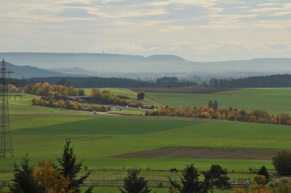 Vom Aussichtsturm bei Villingen geht der Blick nach Süden über die Baar-Hochmulde zur südlichen Baaralb mit Fürstenberg und Länge Von erhöhtem Standort blickt man über eine weite, flachhügelige Landschaft mit Äckern und Wiesen. Mehrere Waldstreifen durchziehen das Gelände. Im Hintergrund sind bewaldete Berge erkennbar.