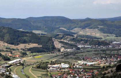 Blick aus großer Höhe auf eine breite Flussaue mit Siedlung. Die Aue wird zum Hintergrund hin von bewaldeten Bergen gesäumt. Etwa in der Bildmitte ist ein Steinbruch zu erkennen.