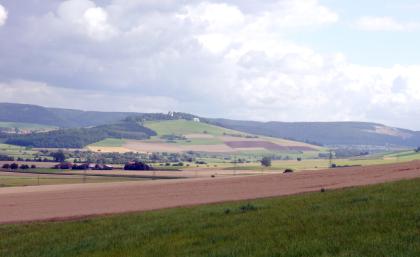 Der Wartenberg von Südwesten Blick über Wiesen und Felder auf gegenüberliegende, teils bewaldete, teils von Äckern durchzogene mittelhohe Berghänge. Ganz rechts befindet sich am dortigen Berghang ein Steinbruch.