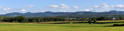 Albtrauf zwischen dem Schönberg bei Pfullingen (links) und dem Rossberg bei Reutlingen-Gönningen (rechts) Panoramabild einer bewaldeten Bergkette mit teils gerundeten Kuppen, teils flach abgestuften Höhen. Davor liegen ausgedehnte Wälder, Maisfelder und sanft ansteigende Wiesen.