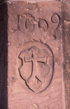 Grenzstein mit Zeichen der Freiburger Münsterbauhütte Das Foto zeigt einen rötlich grauen Grenzstein mit eingraviertem, kreuzähnlichem Zeichen sowie einer Jahreszahl darüber.