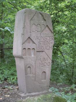 Sandsteinstele oberhalb der nördlichen Brüche Das Foto zeigt einen aufrecht stehenden, leicht rötlich grauen Stein, in den vereinfacht dargestellte Kirchtürme sowie die Worte „Münstersteinbruch um 1200“ eingemeißelt sind.