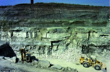 Abbau des Gipslagers der Grundgipsschichten Blick von oben auf eine Steinbruchwand mit grünlichen Gesteinsschichten rechts und dunkler Auflage darüber. Im unteren Bildteil sind Bagger und Radlader zu sehen.