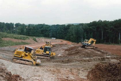 Das Bild zeigt den Abbau in einer Tongrube. Mehrere Raupenfahrzeuge links sowie ein Bagger rechts sind dabei auf einem Hügel im Vordergrund im Einsatz. Ein Waldstreifen im Hintergrund begrenzt das Abbaugelände.