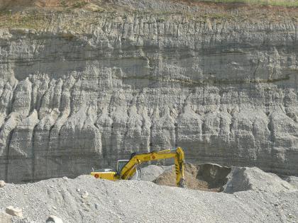 Gewinnung im Trockenabbau in der Kiesgrube Schaffhausen Blick auf eine bildfüllende Steinbruchwand. Dem grau bis bräunlich gefärbten, zahnartig gefurchtem Gestein ist eine Abraumhalde mit Bagger vorgelagert.