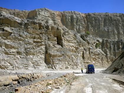 Muschelkalklagerstätten liefern hochwertige Rohstoffe für den Verkehrswege-, Hoch- und Tiefbau Blick in einen Steinbruch mit zwei sehr hohen, gebirgsähnlichen Wänden. Die vordere, bräunlich graue Gesteinswand steigt nach links hin an. Im Vordergrund links Gesteinsreste sowie rechts ein Fahrweg mit abgestelltem Fahrzeug.